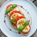 Golden seared chicken breast topped with fresh tomatoes, melted mozzarella, basil and balsamic glaze on white plate