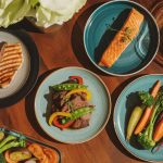 Variety of high protein dinner recipes including grilled chicken, salmon, and beef arranged on a rustic wooden table with weights in background