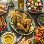 Colorful Mediterranean dinner table with grilled chicken, fresh vegetables, olive oil, feta cheese, and herbs on rustic wooden table