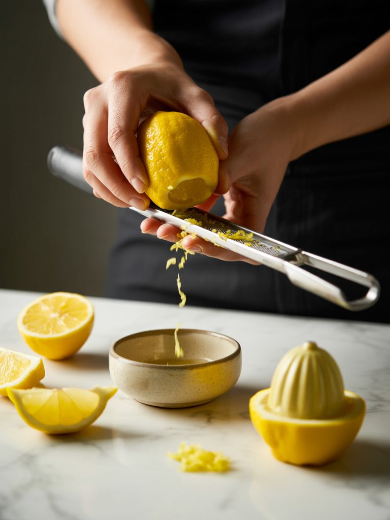Hands zesting bright yellow lemon with microplane grater over bowl with halved lemons nearby
