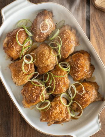 Golden-brown baked teriyaki chicken thighs with caramelized glaze on white platter, garnished with sesame seeds and green onions