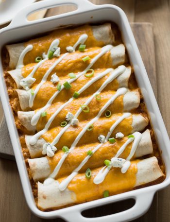 Buffalo chicken enchiladas in a baking dish drizzled with ranch dressing and topped with blue cheese crumbles and green onions