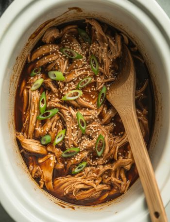 Overhead view of tender shredded teriyaki chicken in slow cooker with glossy sauce and garnish