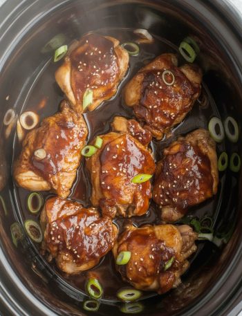 Slow cooker filled with tender teriyaki chicken thighs in glossy sauce, garnished with sesame seeds and green onions