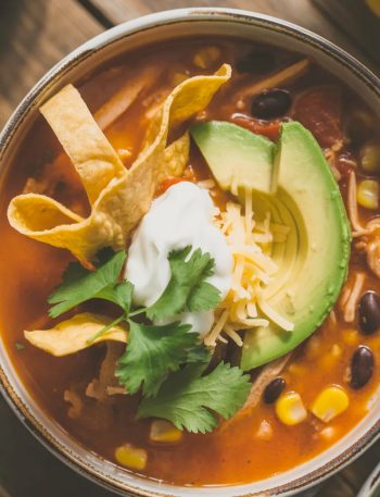 Bowl of classic chicken tortilla soup topped with crispy tortilla strips, fresh avocado slices, sour cream, shredded cheese, and cilantro on a rustic wooden table