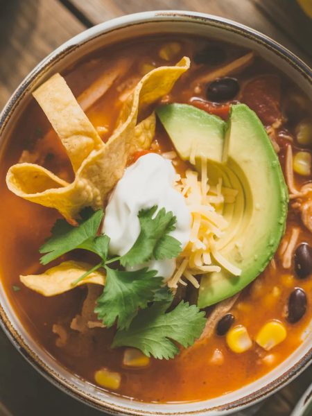 Bowl of classic chicken tortilla soup topped with crispy tortilla strips, fresh avocado slices, sour cream, shredded cheese, and cilantro on a rustic wooden table