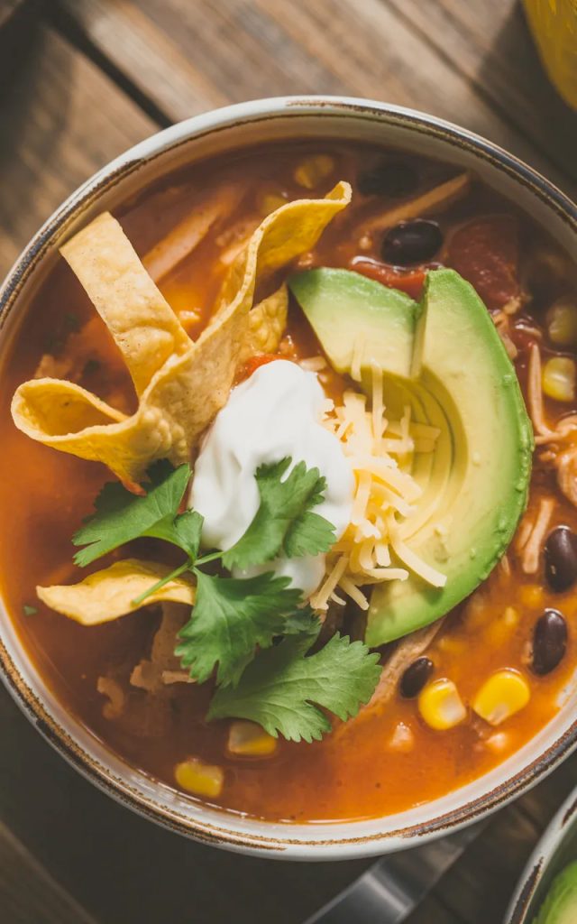 Bowl of classic chicken tortilla soup topped with crispy tortilla strips, fresh avocado slices, sour cream, shredded cheese, and cilantro on a rustic wooden table