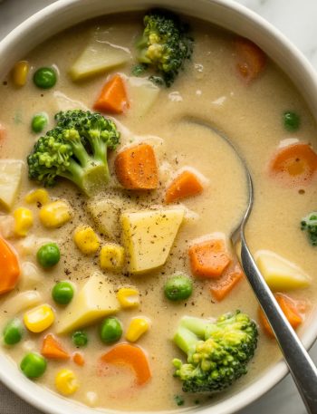 White bowl filled with creamy vegetable soup showing colorful broccoli, carrots, corn, potatoes, and peas in a rich, velvety broth with a spoon