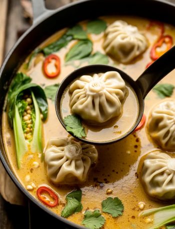 Black pot filled with Thai potsticker soup showing dumplings in creamy coconut broth with fresh cilantro, red chili flakes, and a black ladle