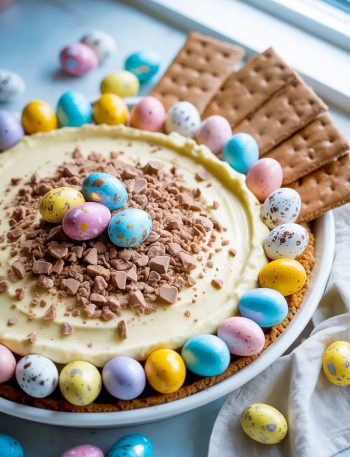 A white ceramic bowl filled with creamy Cadbury eggs dessert dip topped with crushed Cadbury mini eggs, surrounded by colorful pastel blue, yellow, pink, and white whole Cadbury mini eggs arranged around the edge of the bowl, served with chocolate graham crackers on the side, on a light blue surface