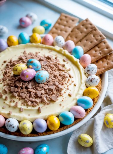 A white ceramic bowl filled with creamy Cadbury eggs dessert dip topped with crushed Cadbury mini eggs, surrounded by colorful pastel blue, yellow, pink, and white whole Cadbury mini eggs arranged around the edge of the bowl, served with chocolate graham crackers on the side, on a light blue surface