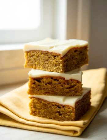 Stack of three moist carrot cake bars with cream cheese swirl on a yellow linen napkin on a white surface