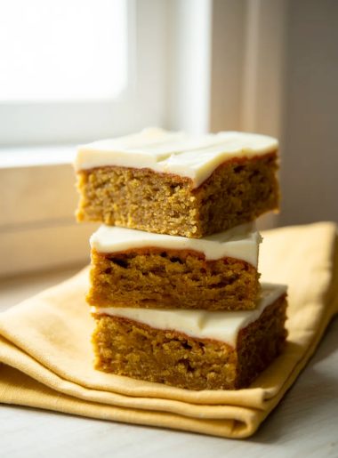 Stack of three moist carrot cake bars with cream cheese swirl on a yellow linen napkin on a white surface