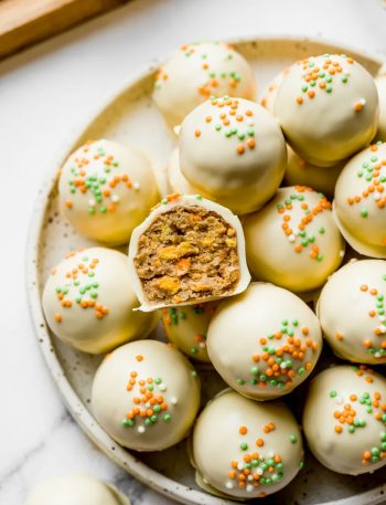Carrot cake truffles on a speckled ceramic plate coated in smooth white chocolate with orange and green nonpareil sprinkles one truffle cut open showing the moist spiced carrot cake interior