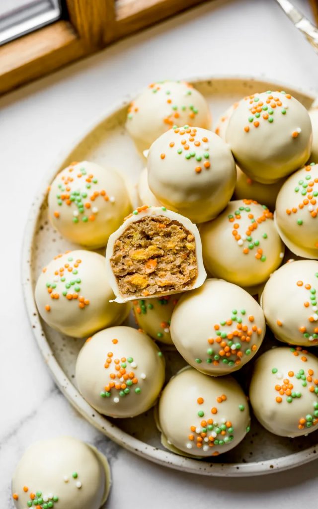 Carrot cake truffles on a speckled ceramic plate coated in smooth white chocolate with orange and green nonpareil sprinkles one truffle cut open showing the moist spiced carrot cake interior