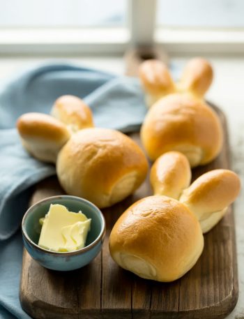Soft golden Easter bunny shaped bread rolls arranged on a rustic wooden board with a small bowl of butter on the side