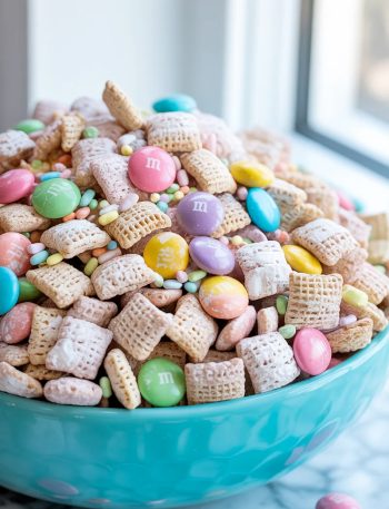 Easter bunny chow made with powdered sugar coated Chex cereal and colorful pastel M&Ms piled high in a turquoise ceramic bowl on a white surface