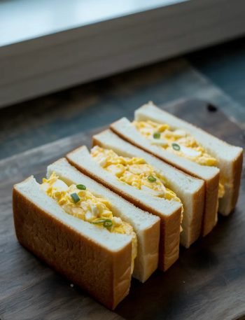 Four rectangular Japanese egg sandwich fingers arranged side by side on a dark wooden surface, each showing a thick cross section of creamy yellow egg salad filling between two slices of soft white crustless milk bread, scattered green onions on top