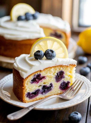 A slice of moist lemon blueberry cake with thick cream cheese frosting garnished with fresh blueberries and a lemon slice on a white decorative plate
