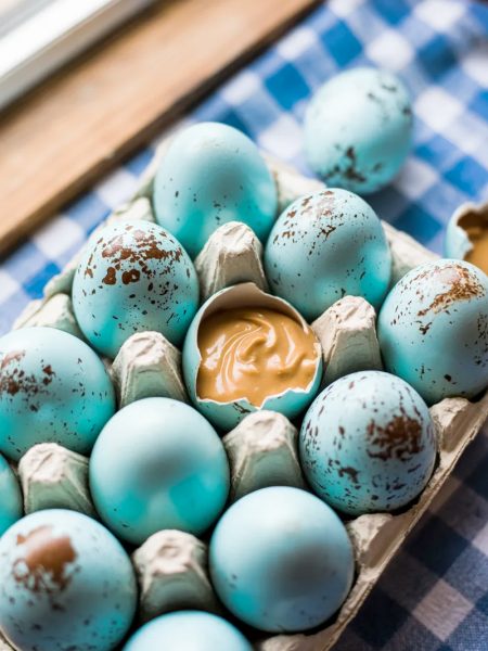 Homemade speckled blue peanut butter Easter eggs in a white ceramic egg carton on a blue gingham tablecloth with one egg broken open showing the creamy peanut butter filling