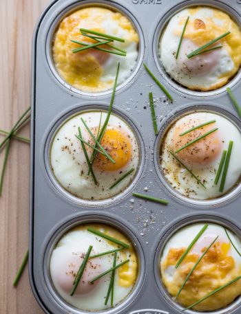 Overhead view of a six cup muffin tin filled with freshly baked French eggs showing silky set egg whites with soft golden cheese tops, fresh sliced chives scattered across each cup, and cracked black pepper on a wooden surface