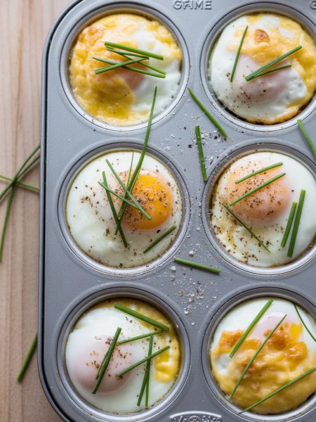 Overhead view of a six cup muffin tin filled with freshly baked French eggs showing silky set egg whites with soft golden cheese tops, fresh sliced chives scattered across each cup, and cracked black pepper on a wooden surface