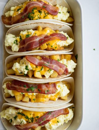 Overhead view of six soft flour tortilla breakfast tacos standing upright side by side in a white rectangular baking dish, filled with fluffy scrambled eggs, crispy bacon, golden home fries, and melted cheddar cheese, garnished with fresh chopped parsley
