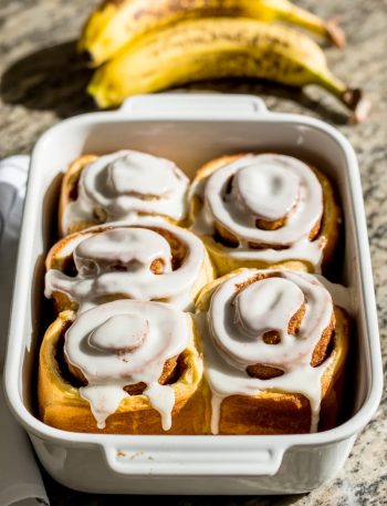 Four golden banana bread cinnamon rolls in a white baking dish covered in a thick vanilla glaze with visible cinnamon swirls and overripe bananas in the background