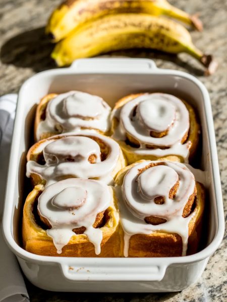Four golden banana bread cinnamon rolls in a white baking dish covered in a thick vanilla glaze with visible cinnamon swirls and overripe bananas in the background