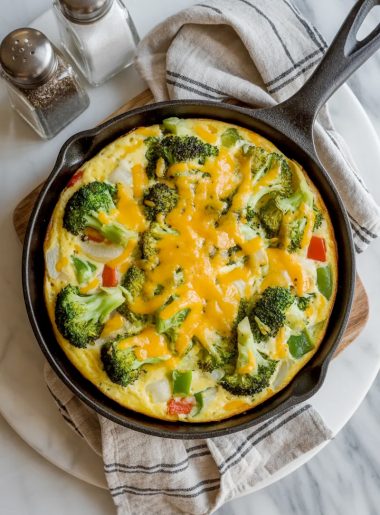 Golden baked vegetable frittata in a black cast iron skillet on a marble surface, loaded with broccoli, red and green bell peppers, and melted cheddar cheese on top
