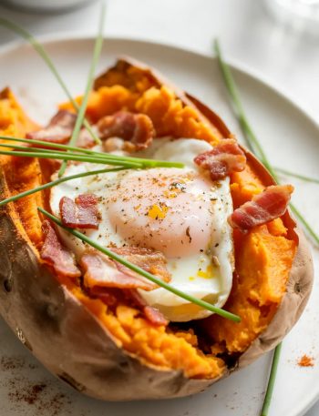 Close up of a baked sweet potato split open with a perfectly set egg nestled in the fluffy orange flesh, crumbled crispy bacon scattered around the egg, fresh chives and cracked black pepper on top, served on a white ceramic plate