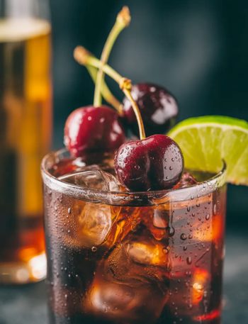 A rocks glass filled with cherry Coke margarita with amaretto over ice garnished with fresh dark cherries and a lime wedge on a dark moody surface