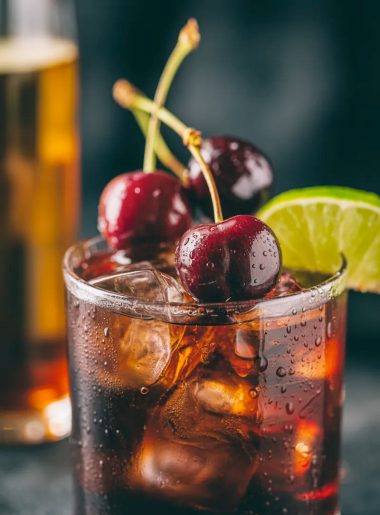 A rocks glass filled with cherry Coke margarita with amaretto over ice garnished with fresh dark cherries and a lime wedge on a dark moody surface