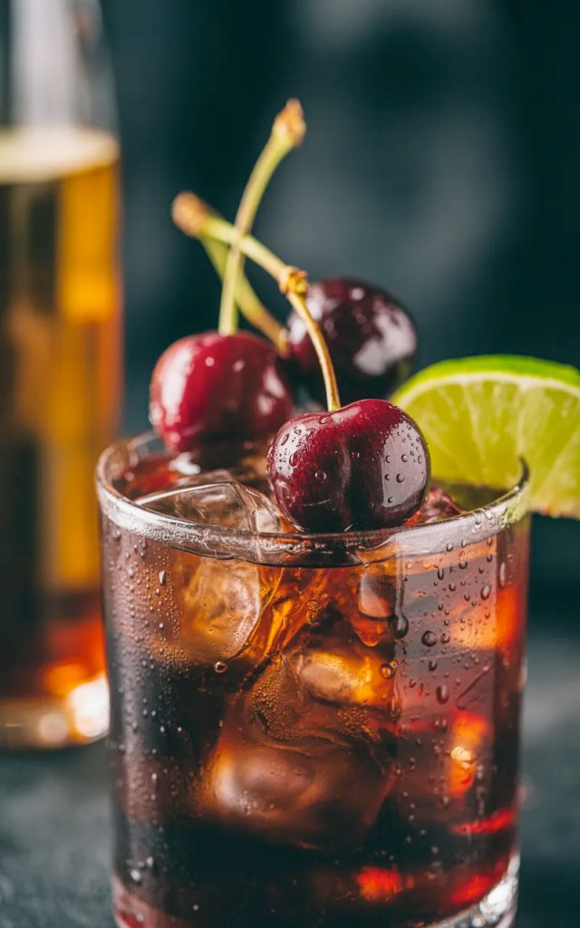 A rocks glass filled with cherry Coke margarita with amaretto over ice garnished with fresh dark cherries and a lime wedge on a dark moody surface