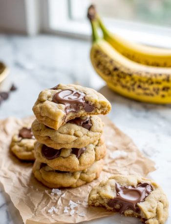 A pile of chewy banana bread chocolate chip cookies on parchment paper with one cookie showing a bite taken out revealing melty chocolate and soft chewy interior with flaky sea salt on top