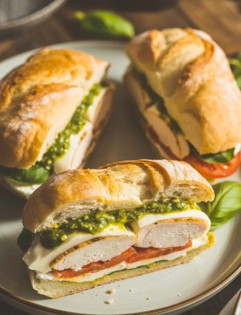 Three golden toasted ciabatta chicken pesto sandwiches on a light grey ceramic plate showing layers of juicy chicken fresh mozzarella basil pesto and ripe tomato