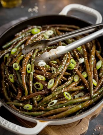 A black cast iron skillet of glossy garlicky Chinese buffet style green beans coated in savory sauce and topped with sesame seeds and sliced green onions