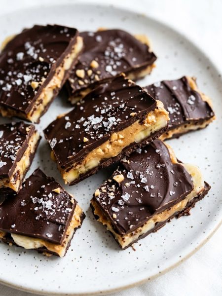 Overhead close-up of broken chocolate banana bark pieces showing peanut butter and banana layers on a white speckled plate