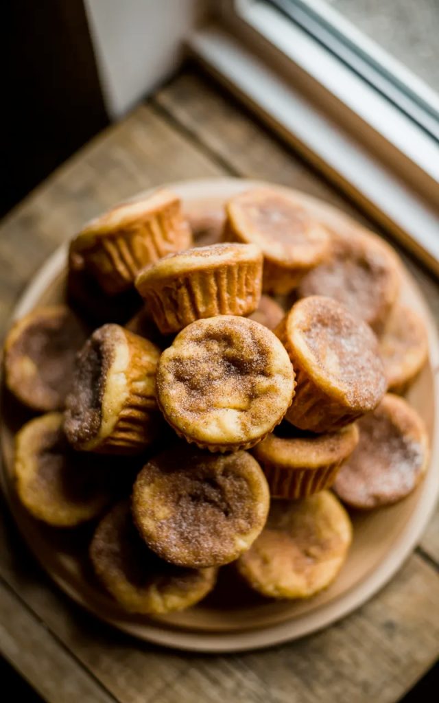 A pile of golden cinnamon sugar French toast muffins with crispy coated exteriors stacked on a white ceramic plate on a wooden surface