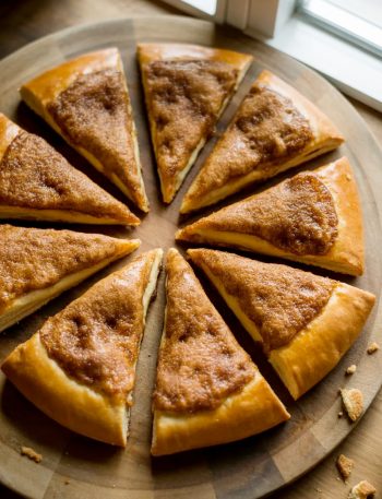 A golden cinnamon sugar crescent roll pizza cut into eight triangular slices arranged on a round wooden board with a caramelized cinnamon sugar crust on top