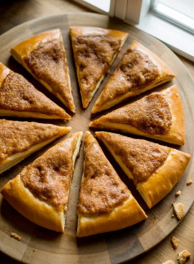 A golden cinnamon sugar crescent roll pizza cut into eight triangular slices arranged on a round wooden board with a caramelized cinnamon sugar crust on top
