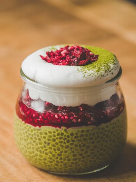 Close-up of a single glass jar of matcha and raspberry chia pudding with three distinct vibrant layers of green matcha chia base, red raspberry compote, and white coconut cream topped with freeze-dried raspberries and matcha dusting
