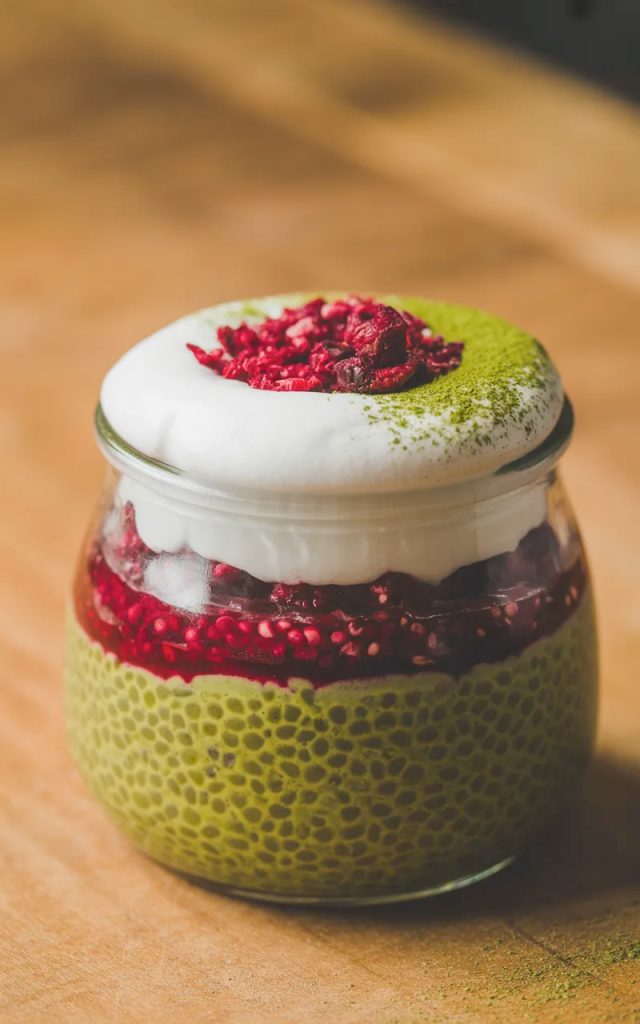 Close-up of a single glass jar of matcha and raspberry chia pudding with three distinct vibrant layers of green matcha chia base, red raspberry compote, and white coconut cream topped with freeze-dried raspberries and matcha dusting