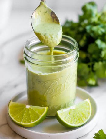 Close-up of a glass jar of creamy cilantro garlic sauce with a spoon lifting a generous scoop of the smooth pale green sauce with lime wedges beside it