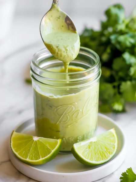 Close-up of a glass jar of creamy cilantro garlic sauce with a spoon lifting a generous scoop of the smooth pale green sauce with lime wedges beside it