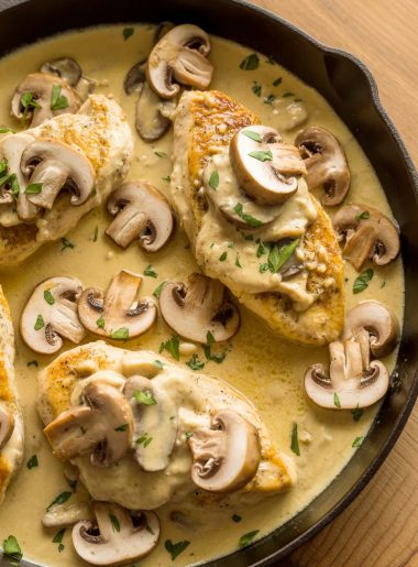 Close-up overhead shot of golden chicken breasts in a rich creamy Asiago mushroom sauce with sliced mushrooms and fresh parsley in a dark skillet on a wooden table