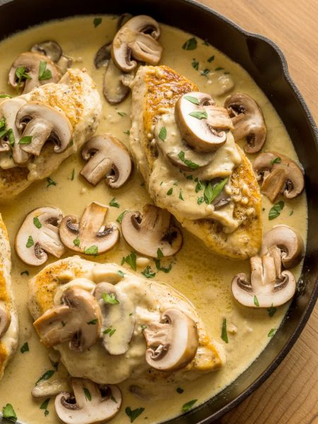 Close-up overhead shot of golden chicken breasts in a rich creamy Asiago mushroom sauce with sliced mushrooms and fresh parsley in a dark skillet on a wooden table