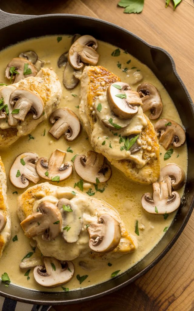 Close-up overhead shot of golden chicken breasts in a rich creamy Asiago mushroom sauce with sliced mushrooms and fresh parsley in a dark skillet on a wooden table