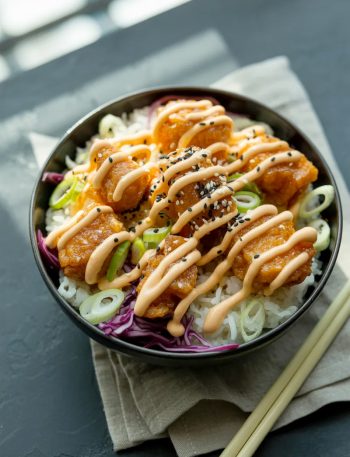 Crispy golden salmon bites piled over white jasmine rice in a dark ceramic bowl drizzled with sriracha mayo and topped with sesame seeds and green onions