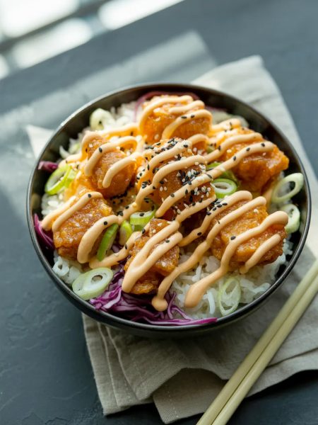 Crispy golden salmon bites piled over white jasmine rice in a dark ceramic bowl drizzled with sriracha mayo and topped with sesame seeds and green onions